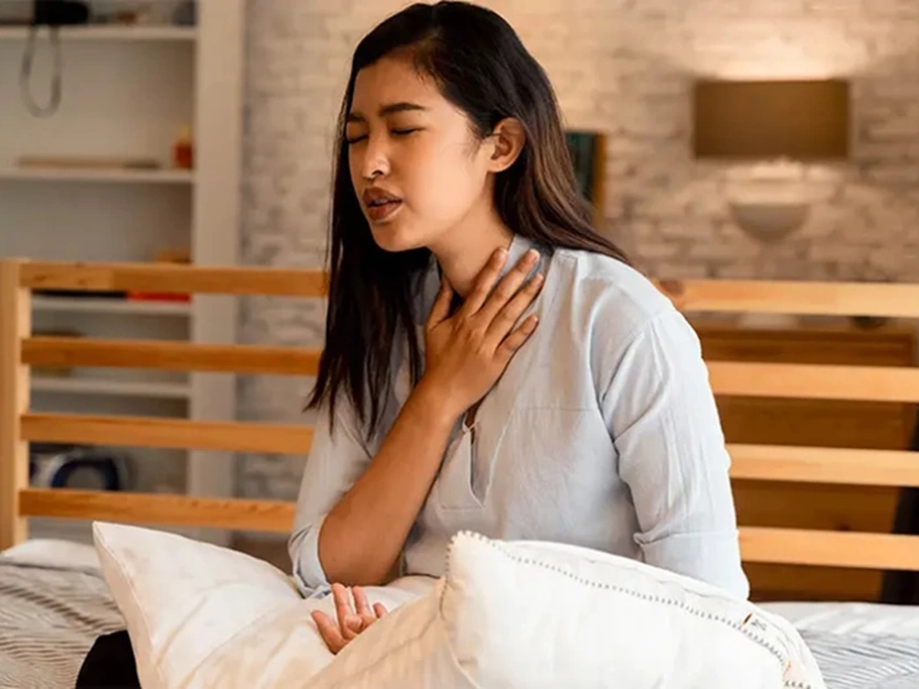 Jeune femme assise sur un lit se tenant la gorge en raison d’un mal de gorge ou d’une gêne respiratoire.